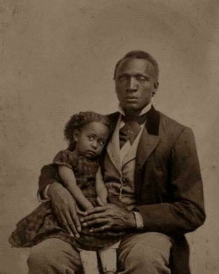 Historical photograph of a man in formal attire holding a young girl on his lap, both looking at the camera.