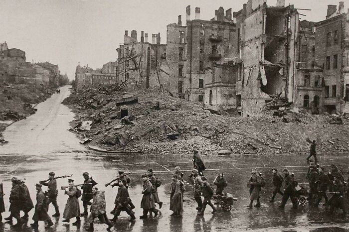 Historic scene of soldiers marching past ruined buildings in a WWII-era city, offering a rare glimpse not in textbooks.