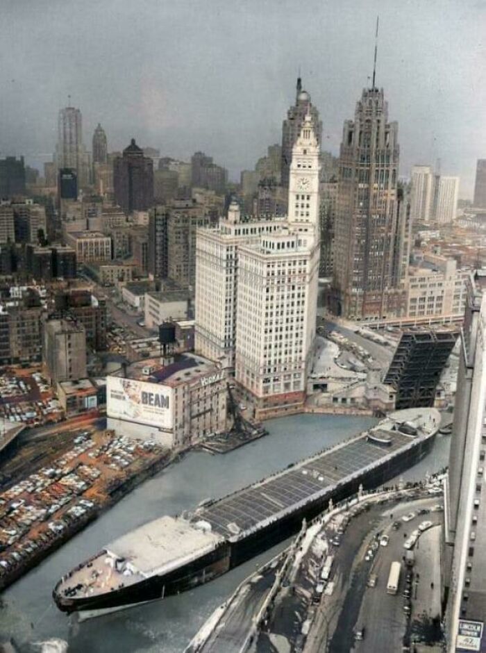 Historic photo of a large ship navigating through a city's river amidst towering skyscrapers.