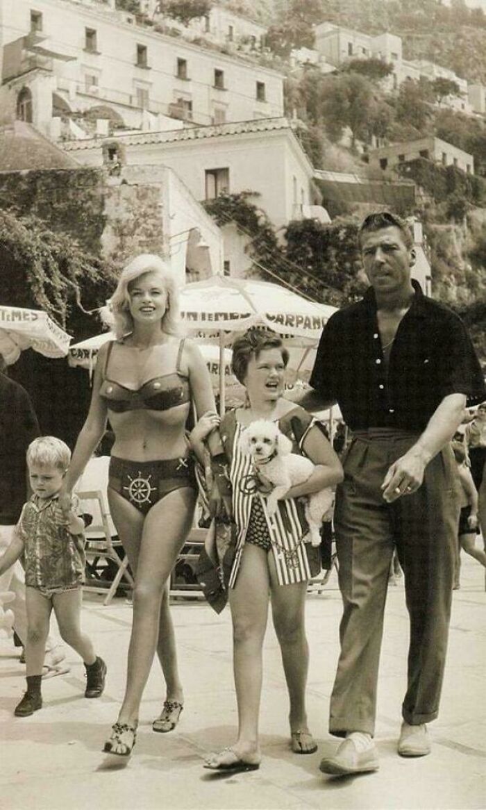 Vintage beach scene with a woman in a bikini, two children, and a man strolling in a historic setting.