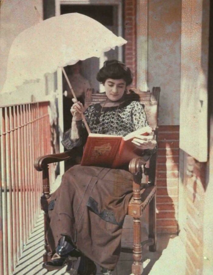 A woman in vintage attire reads on a porch, shielded by a parasol.