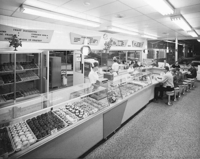 Vintage donut shop interior, with customers seated at a counter and staff behind display cases filled with pastries.