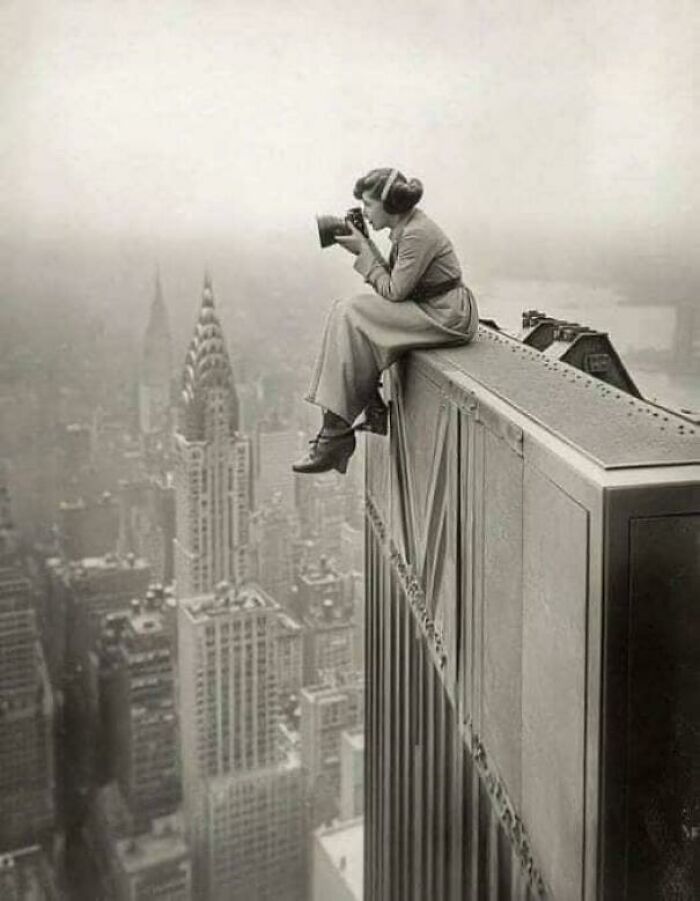 Historic photo of a woman with a camera perched on a skyscraper edge, capturing a breathtaking cityscape view.