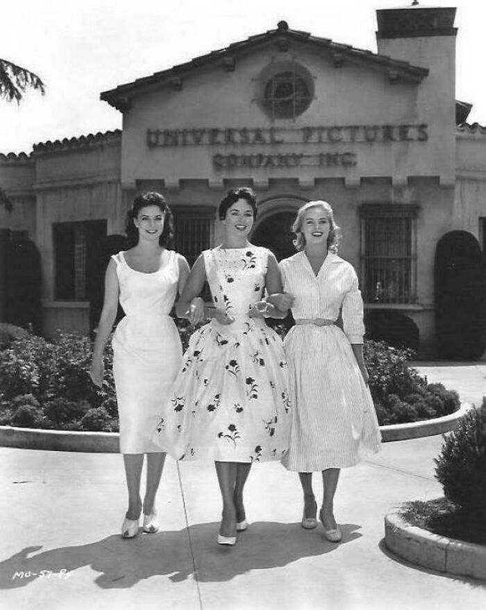 Three women in vintage dresses walking arm-in-arm outside Universal Pictures building, historical photo.