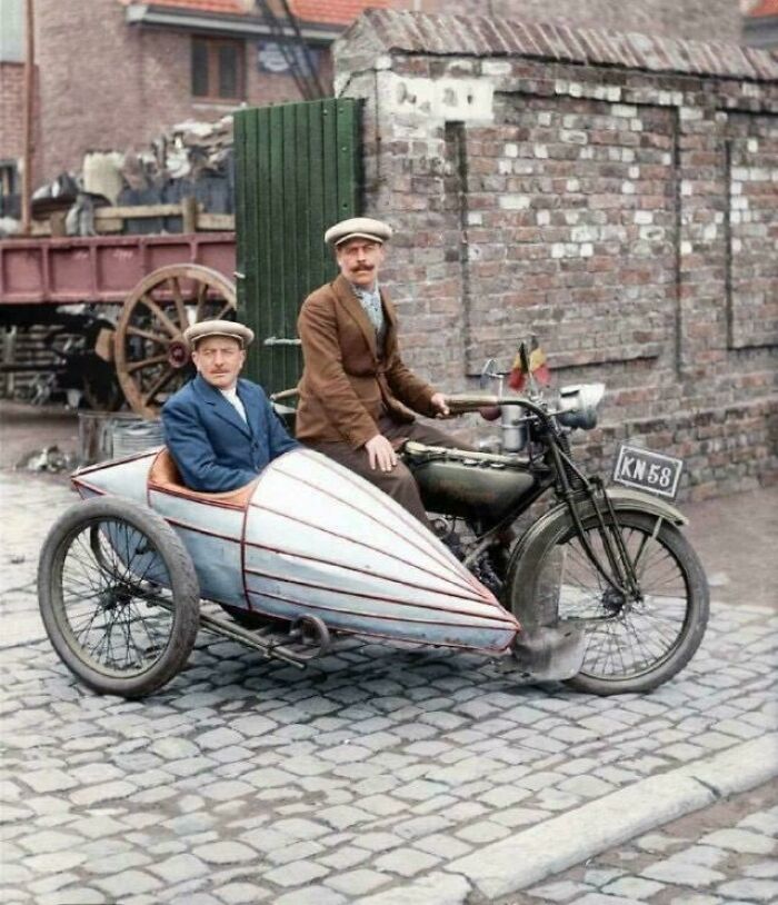 Vintage motorcycle with a sidecar carrying two men in historical attire on a cobblestone street.