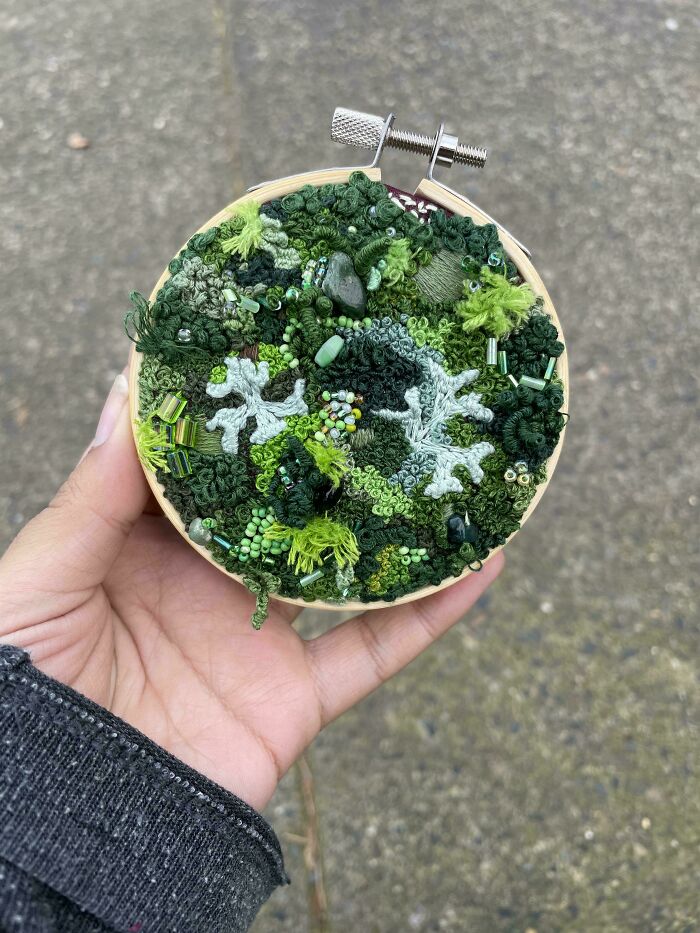 Hand holding a small hoop with textured green embroidery designs featuring beads and various stitches.