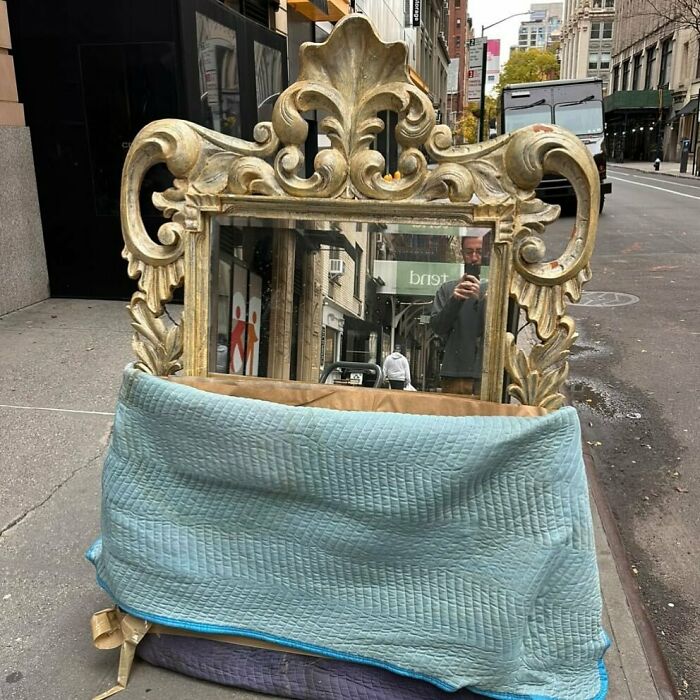 Ornate mirror with gold frame on NYC curb, covered partially by a blanket, reflecting the busy street.