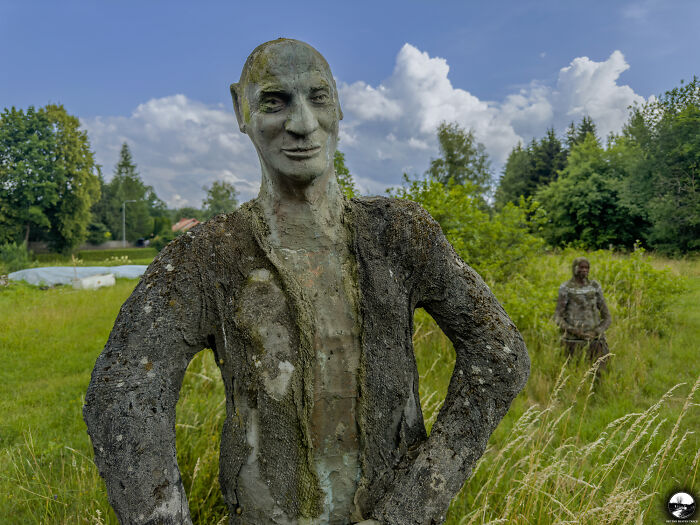 Stone sculpture by Adam Szubski standing in a lush field under a cloudy sky, with another sculpture in the background.
