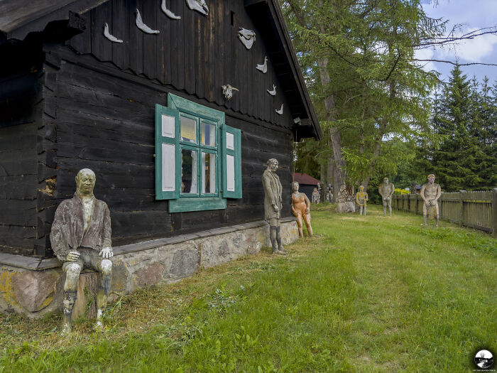Sculptures by Adam Szubski outside a rustic house, set in a grassy area with trees in the background.
