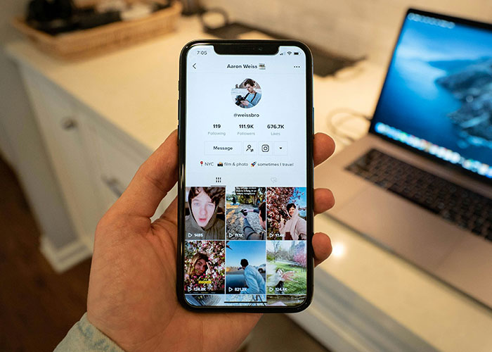 Hand holding a smartphone showing a social media profile, with background of a laptop on a desk; captures '90s and '00s digital trends.