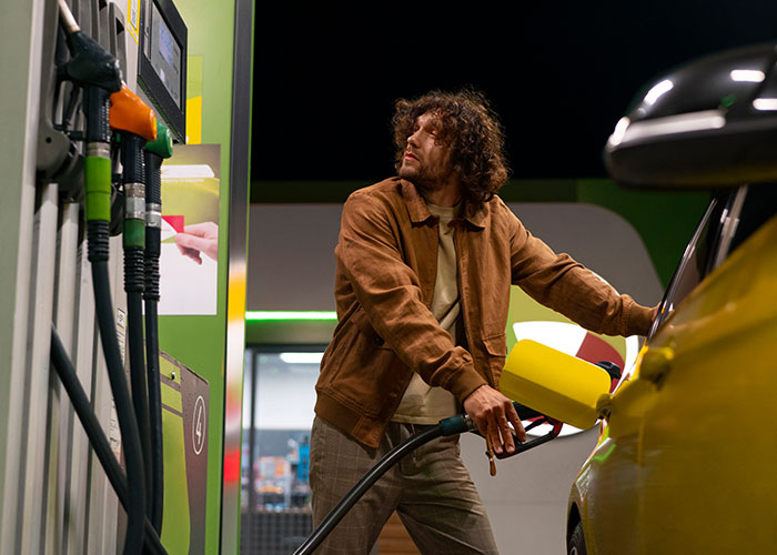 Person in a brown jacket refueling a yellow car at a gas station, evoking nostalgia for '90s and '00s trends.