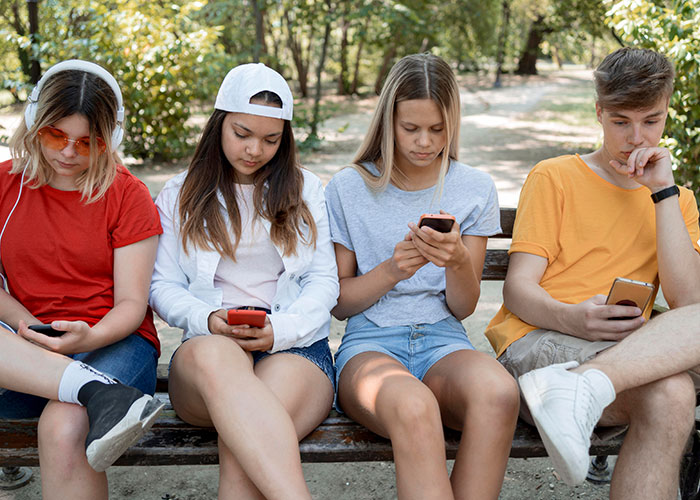 Teens on a bench, absorbed in their phones, embodying trends of the '90s and '00s fashion with casual summer outfits.