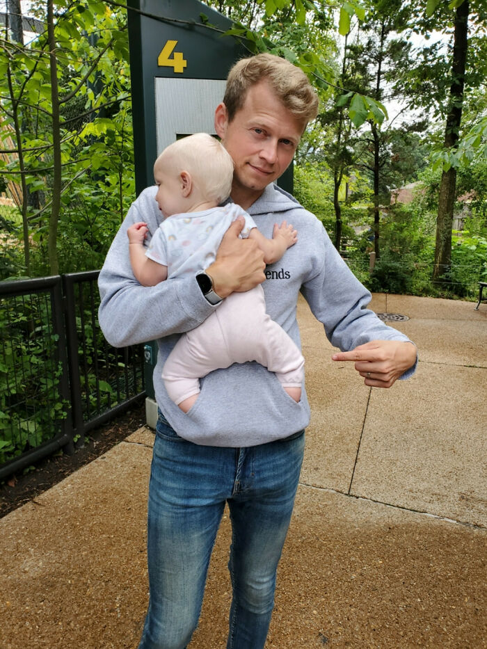 Man holding a baby in a clever way, demonstrating parenting genius tricks, outdoors on a path with greenery.