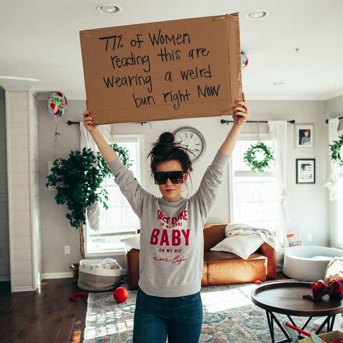 Woman holding a sign about hairstyles, wearing sunglasses, in a cozy living room setting.