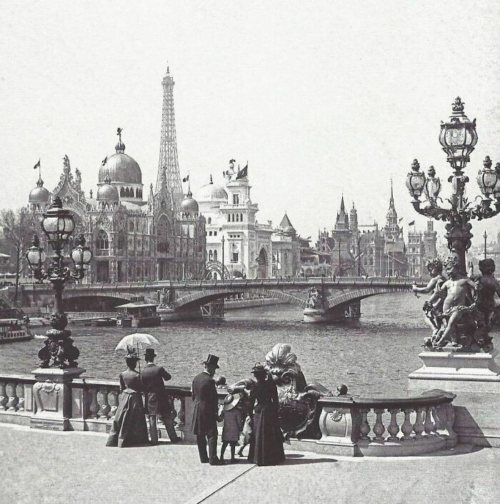 Historical photo of a 19th-century world fair with people in formal attire admiring ornate buildings and a bridge.