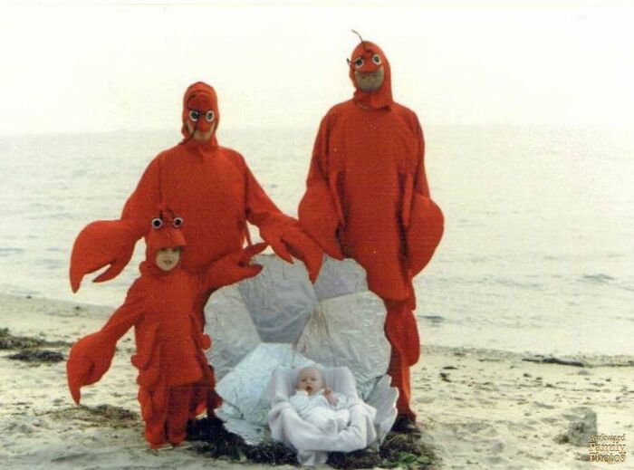 Family photo of three people dressed as lobsters with a baby on a beach.