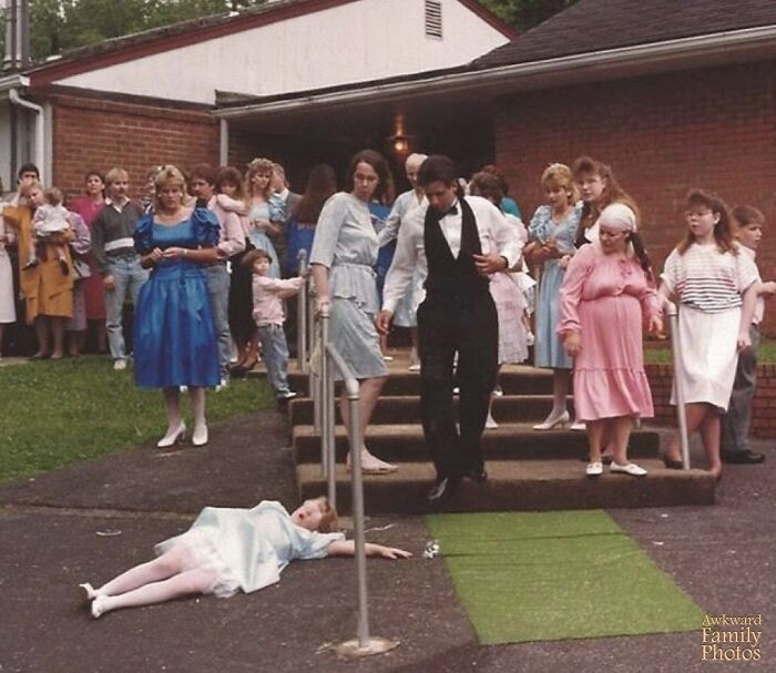 Awkward family photo with a girl dramatically lying on the ground during a formal gathering, surrounded by surprised people.