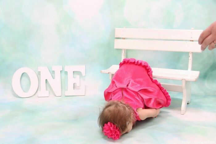 Baby in a pink dress faceplants in front of a white bench and "ONE" sign, creating a viral family photo moment.