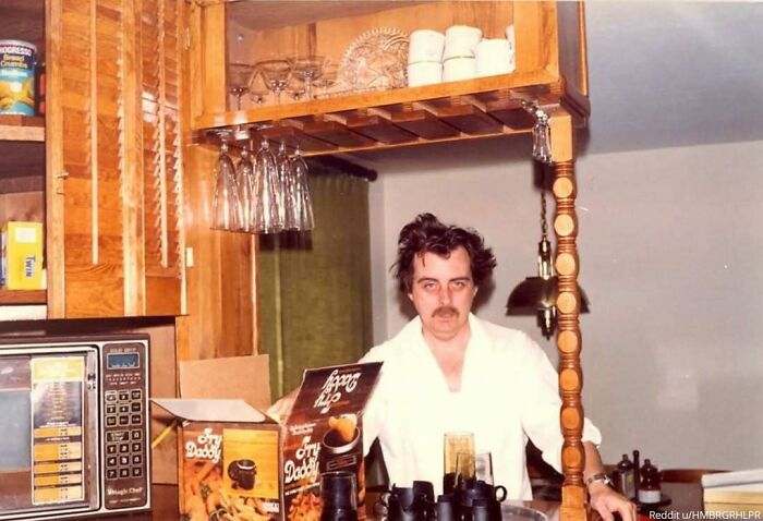 Man with messy hair standing in a cluttered kitchen with wood cabinets and kitchenware, capturing a memorable family photo.