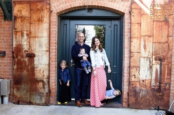 Awkward family photo of parents with children, one child peeking from behind the mother, standing in front of rustic doors.
