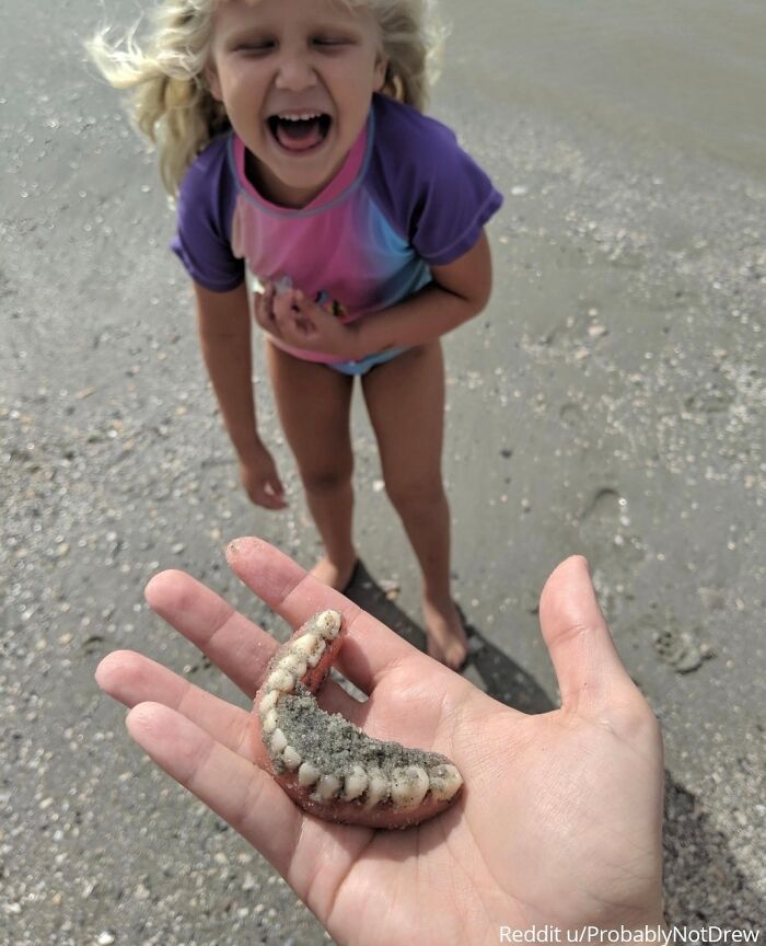 Child laughing on beach while someone holds a set of dentures covered in sand; a viral family photo memory.