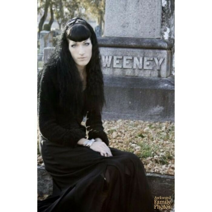 Person in black attire sitting by a gravestone in an awkward family photo setting.
