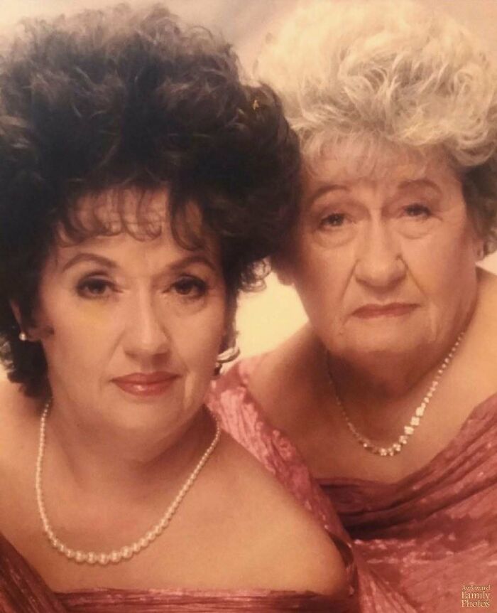 Two women in matching pink dresses with curly hair and pearl necklaces, posing for an awkward family photo.