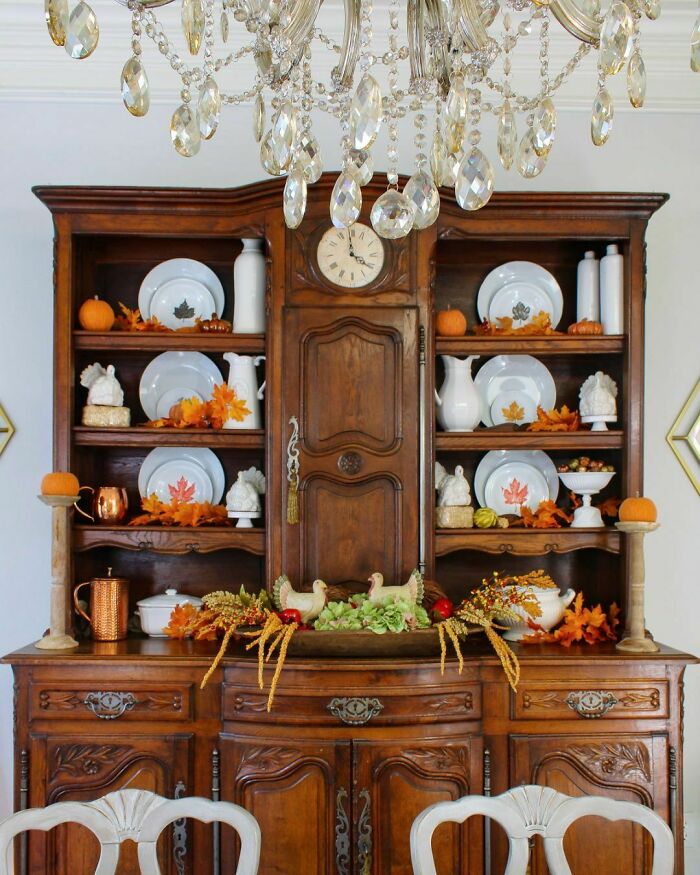 Thanksgiving home decorations on a wooden cabinet with pumpkins, leaves, and ceramic dishes under a crystal chandelier.