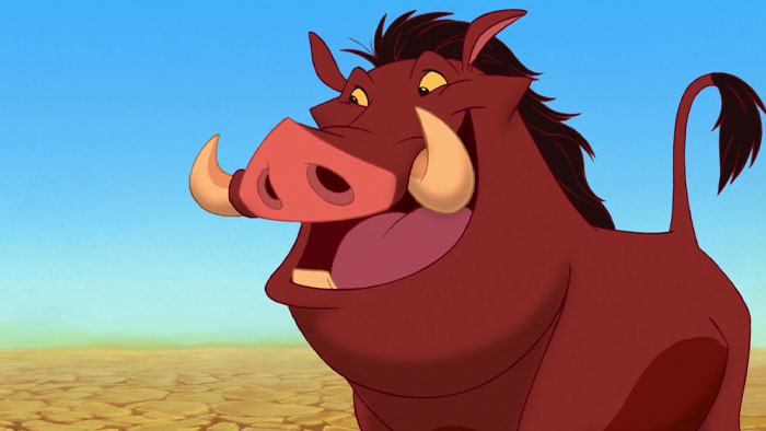 A fat cartoon character, Pumbaa the warthog, smiling widely with prominent tusks, stands on a dry, c*****d landscape under a clear blue sky.