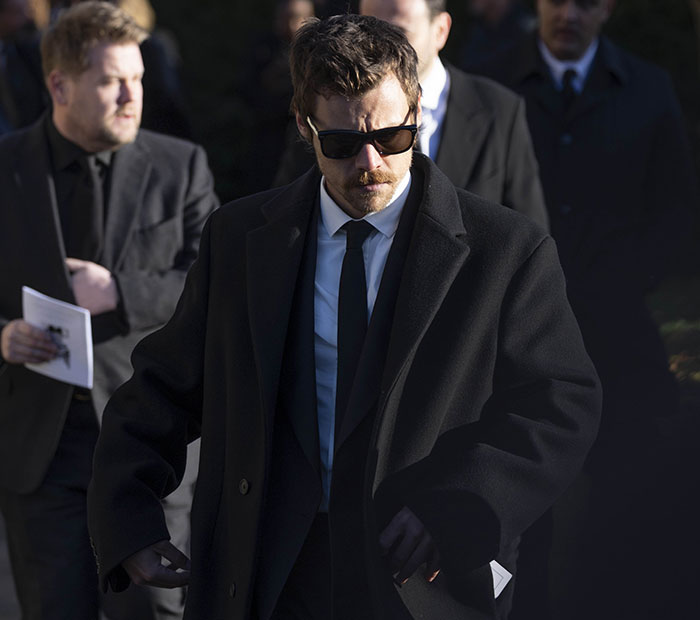 Individual in a black coat and sunglasses, walking solemnly at a formal event. Individual in a black coat and sunglasses, walking solemnly at a formal event.
