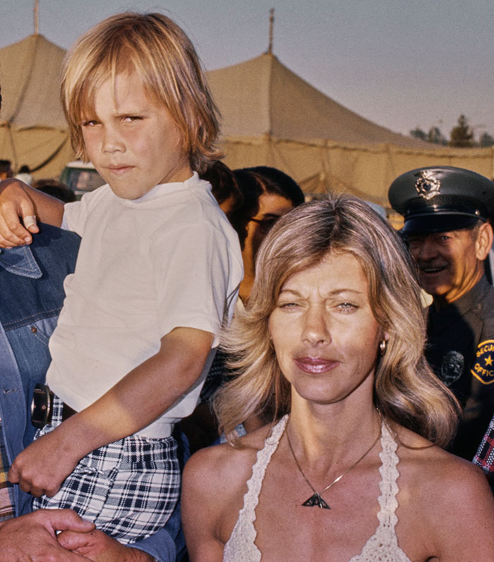 Mother and child outdoors, woman with long blonde hair and boy in white shirt, standing in front of tents and a police officer.