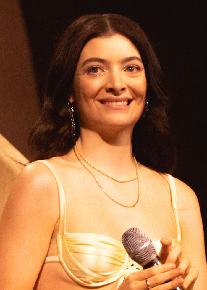 A woman in a light outfit holding a microphone, smiling on stage, related to conspiracy theories discussion.
