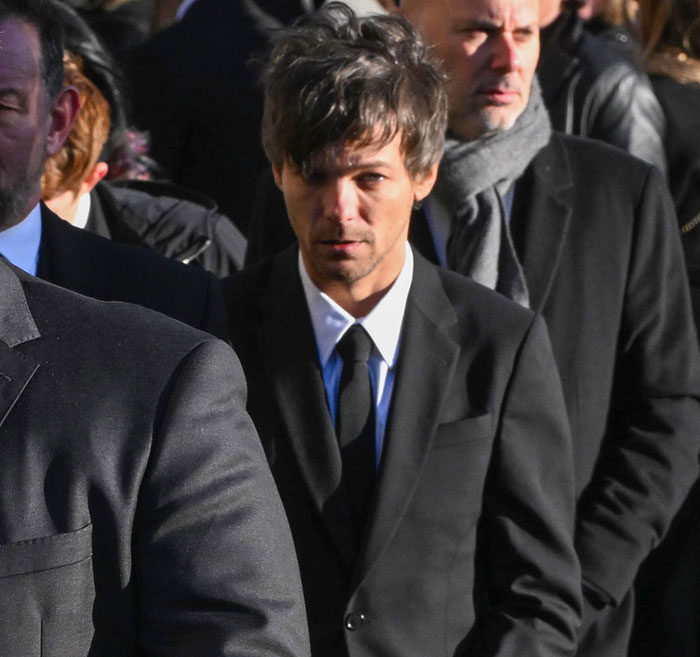 A man in a black suit and tie, looking serious, stands among a crowd, capturing a somber moment. A man in a black suit and tie, looking serious, stands among a crowd, capturing a somber moment.