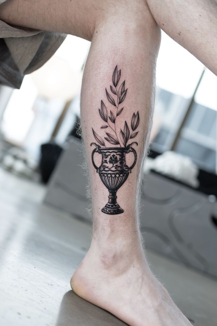 Tattoo of a decorative vase with leaves on a person's leg, created by Monolith Studio artists.