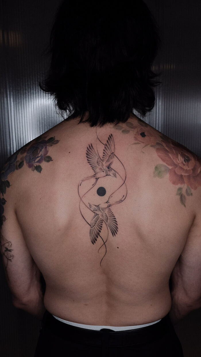 Back tattoo of two birds and a circle, surrounded by colorful floral designs by Monolith Studio artists.