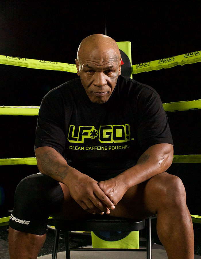 Mike Tyson sitting in a boxing ring, wearing a black shirt, with yellow ropes in the background. Mike Tyson sitting in a boxing ring, wearing a black shirt, with yellow ropes in the background.