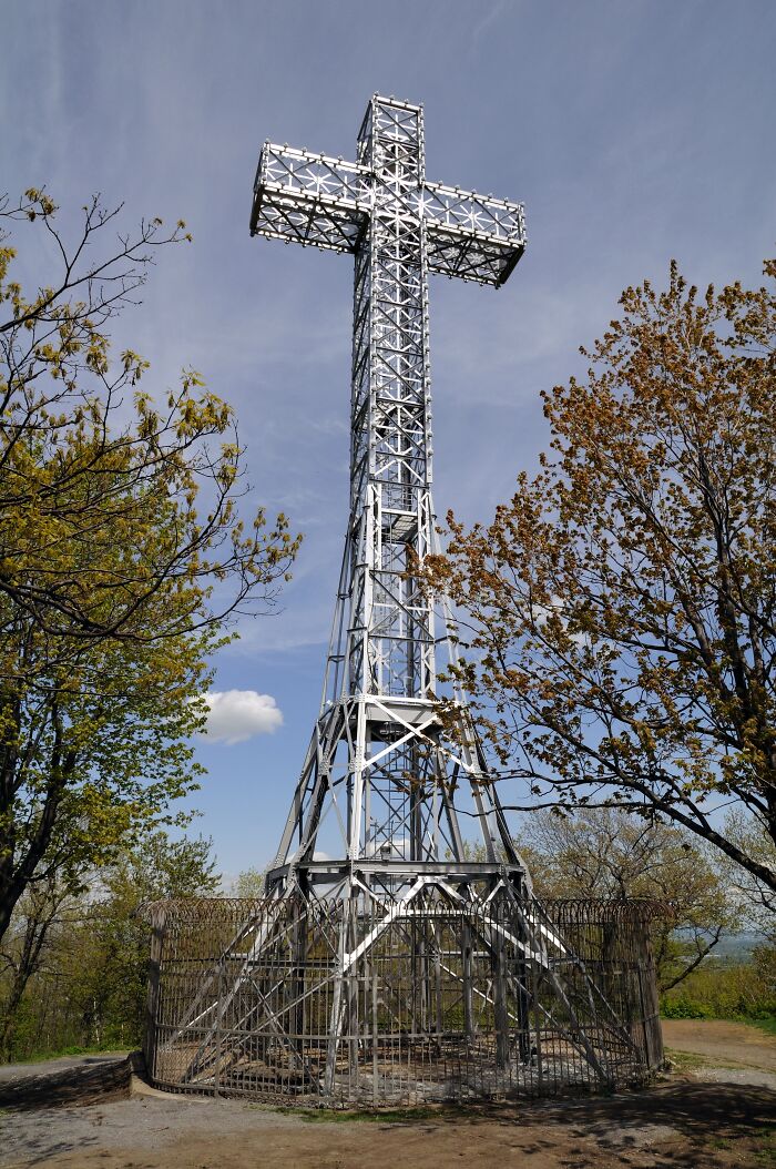 Metallic cross structure, a historical monument surrounded by trees, under a clear blue sky.