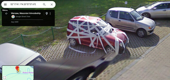 A small car wrapped in white tape, captured on Google Street View in a parking lot.