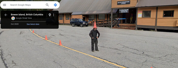 Person with a traffic cone on their head, standing on a street in front of a lodge, captured by Google Street View.