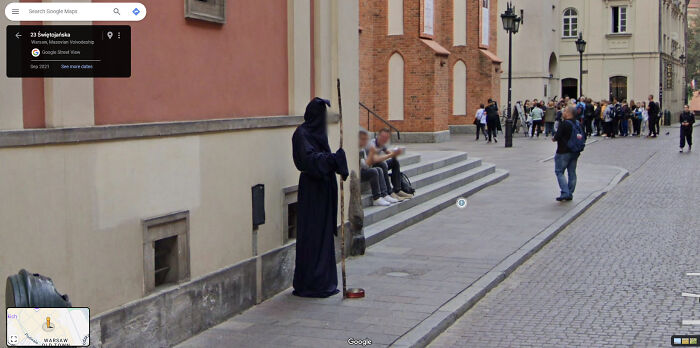 Google Street View captures a person in a hooded costume leaning against a building with people walking by.
