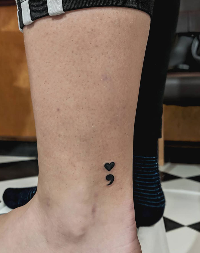 Semicolon tattoo on lower leg with a heart, symbolizing love and resilience against a casual attire background.