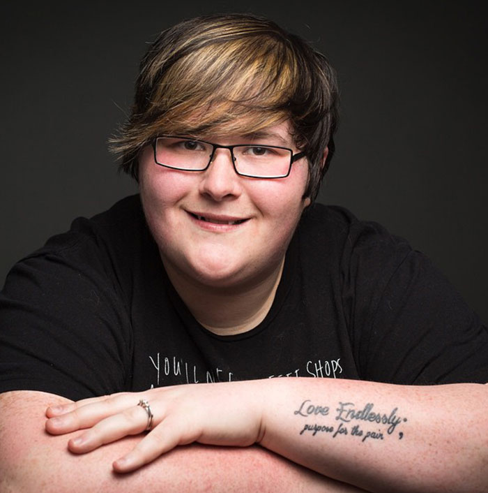 A person with glasses smiling, displaying a semicolon tattoo on their forearm that reads, 