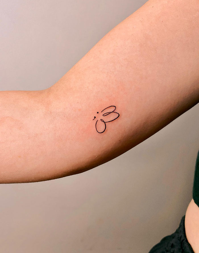 Semicolon tattoo as a minimalist butterfly on inner arm, symbolizing transformation and resilience against a light background.