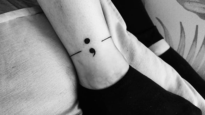Semicolon tattoo on an ankle, integrated into a minimalist line band, symbolizing strength and continuity.