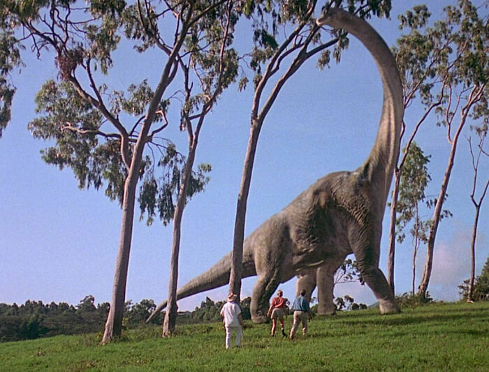 A large dinosaur walking among tall trees, observed by three people in a lush green landscape.