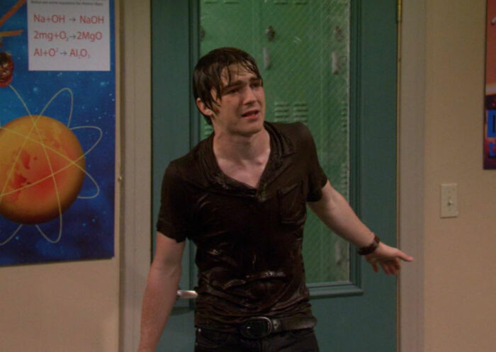 Wet individual standing indoors, dressed in a dark shirt, in front of a science-themed poster.