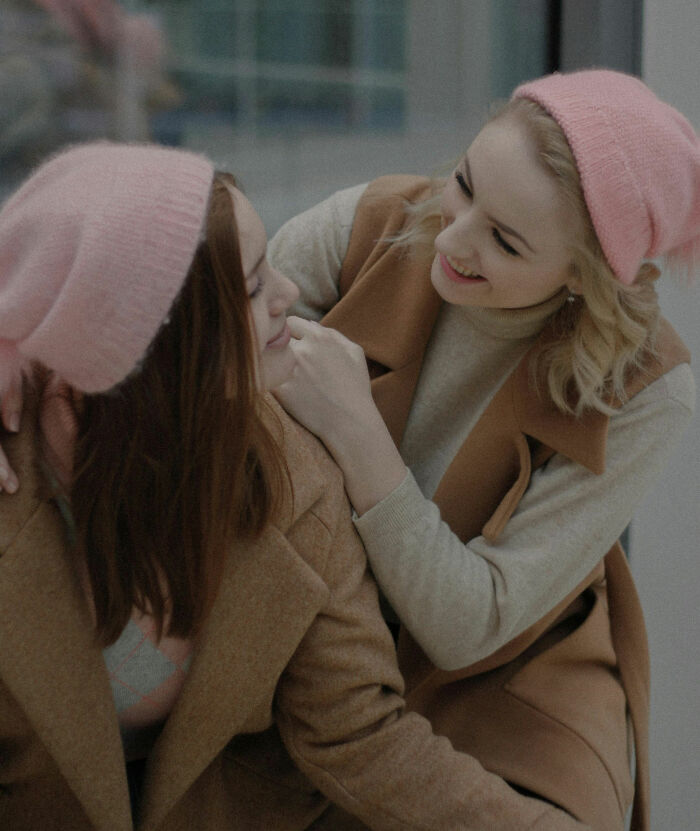 Two women in pink hats and brown coats smiling at each other, illustrating the theme of questioning assumptions.