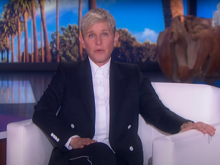 Talk show host sitting on a white couch, wearing a black suit, with a California landscape backdrop.