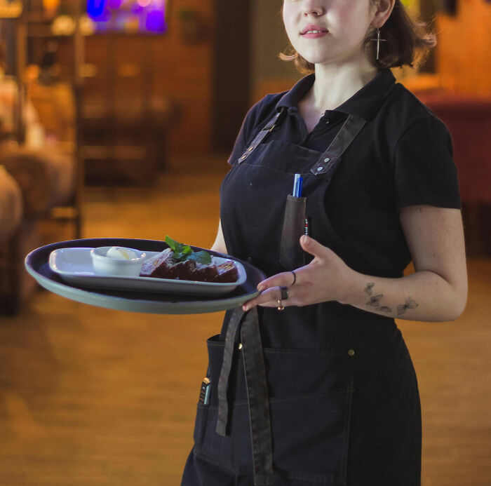 A server in a black apron holding a tray of dessert in a restaurant setting.