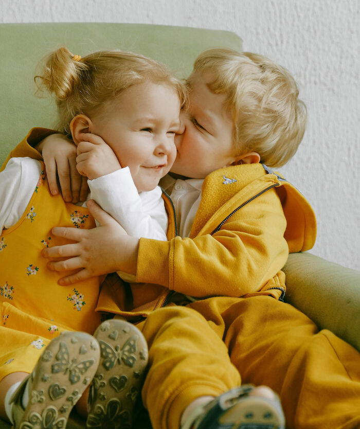 Two children sitting on a green chair, one kissing the other, both in matching yellow outfits, highlighting innocence.